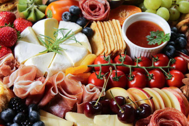Image of charcuterie board, soft and hard cheeses, tomatoes, strawberries, ham and salami roses, rows of crackers, kiwi, vine tomatoes, blueberries, figs, ramekins of chutney, peppers, focus on foreground