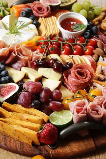 Image of charcuterie board, soft and hard cheeses, tomatoes, strawberries, ham and salami roses, rows of crackers, kiwi, vine tomatoes, blueberries, figs, ramekins of chutney, lime, focus on foreground