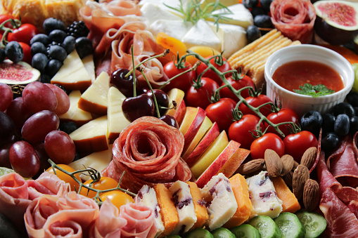 Stock photo showing close-up view of wooden charcuterie board covered with prepared sliced and chopped ingredients including soft and hard cheeses, tomatoes, strawberries, ham and salami roses, rows of crackers, kiwi, vine tomatoes, blueberries, figs, ramekins of chutney, grapes, olives and cherries.