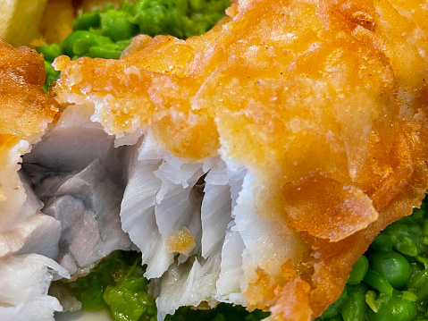 Stock photo showing close-up view of battered cod and chips with mushy peas on a white plate. The fillet of fish is torn open to display the white flesh.
