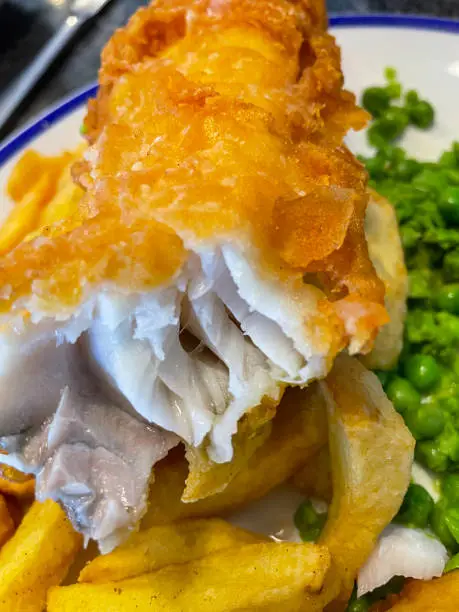 Photo of Close-up image of blue rimmed plate containing flakey white fish and chips, battered cod and French fries served with mushy peas, elevated view, focus on foreground