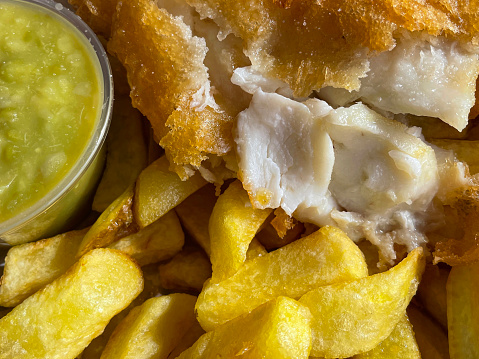 Stock photo showing close-up, elevated view of battered cod and chips with plastic, single-use, disposable container of mushy peas on a white plate. The fillet of fish is torn open to display the white flesh.