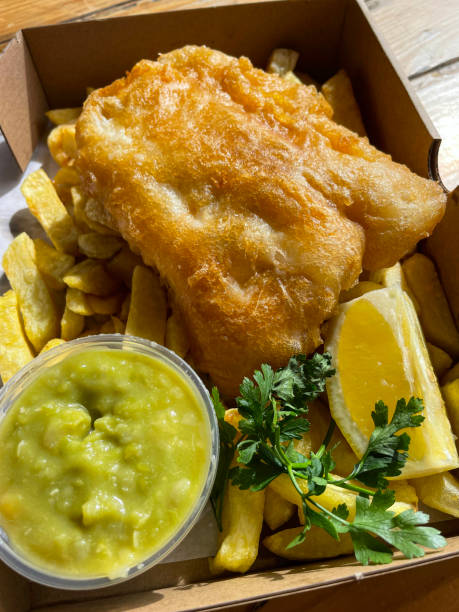istock Close-up image of flakey white fish and chips served in a brown cardboard box, battered cod and French fries, plastic, single-use, disposable container of mushy peas, lemon wedge, elevated view 1440910407