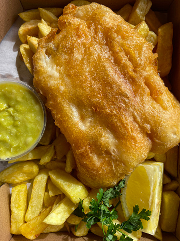 Stock photo showing close-up, elevated view of battered cod and chips with plastic, single-use, disposable container of mushy peas in a brown cardboard box.