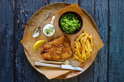 Stock photo showing close-up, elevated view of a wooden platter lined with greaseproof paper that is filled with a portion of battered cod and chips, with a lemon slice, mushy peas and tartare sauce, against a blue wood grain background.