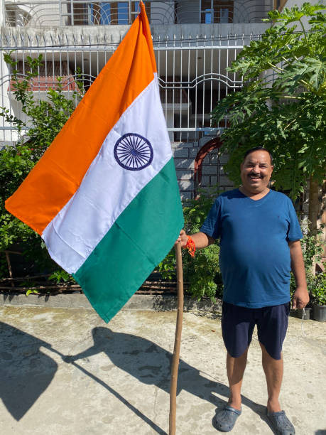 Stock photo showing a residential display of the National flag of India (tricolour) flying from wooden flagpole to celebrate Independence Day. Legally the flag can only be made of silk or khadi cloth. imagen de un hombre indio sosteniendo la bandera nacional de la india (tricolor) volando desde el asta de madera para celebrar el día de la independencia, bandera de tela khadi de azafrán (naranja), rayas blancas y verdes con ashoka chakra azul marino ce