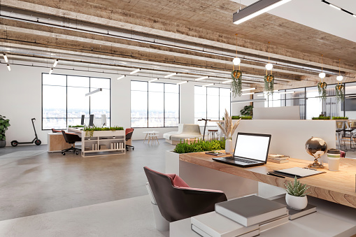 Modern open plan office space interior. Desk, laptop, armchair, vase, plant, concrete floor and ceiling, and large windows in the background. Template for copy space. Render.