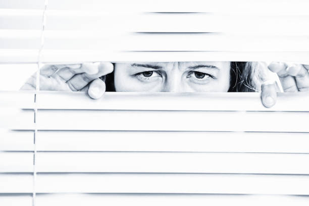 istock Young woman peering anxiously through blinds 1440923045