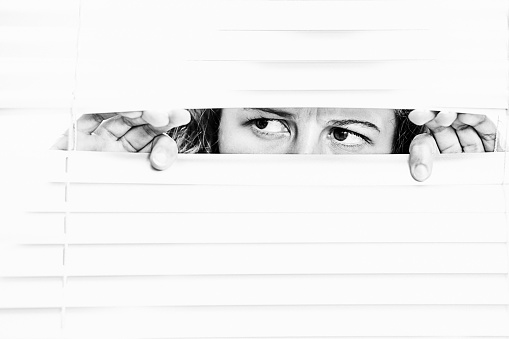 Woman peeking out through window blinds, looking fearful.