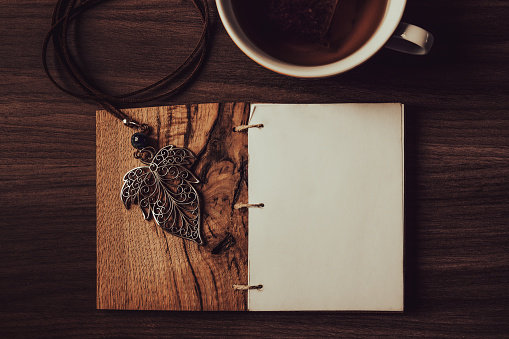 Blank diary page with leaf pendant and cup of tea on wooden background
