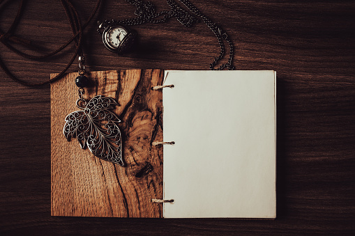 Blank diary page with leaf pendant and pocket watch on wooden background, flat lay