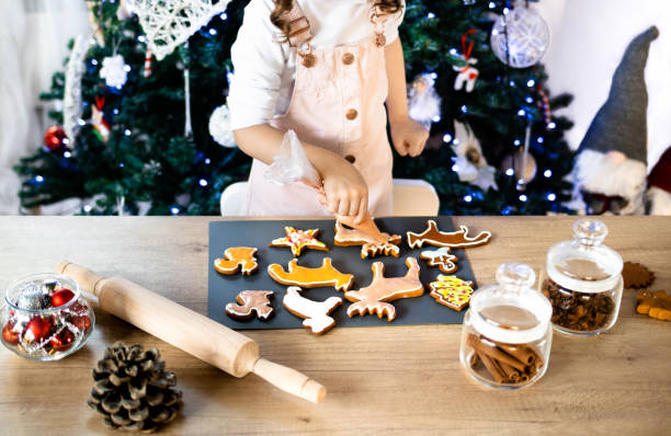 happy little girl coloring christmas gingerbread cookies at home. christmas decoration in the kitchen. fir tree with fairy lights. the concept of the new year and christmas - gift orange green package imagens e fotografias de stock