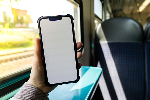 Female hand holding white screen phone in the train. Social Network. Work and Travel phone with blank copy space screen for your text message. New Apps train railway. Phone User. Smartphone with white Screen Mock Up Display