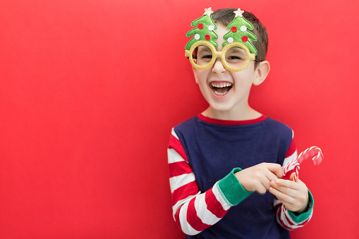 Caucasian boy in funny glasses holding tasty candy canes on a red background, copy space