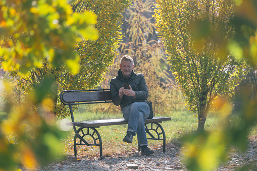 Man is using mobile phone outdoors, sitting on bench at public park