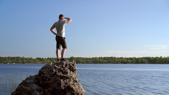 A young man stands on a stone near the river admiring the landscape. A man in nature. 4k