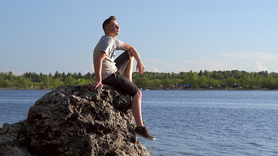 A young man sits on a stone by the river and admires nature. A man in nature closeup. 4k