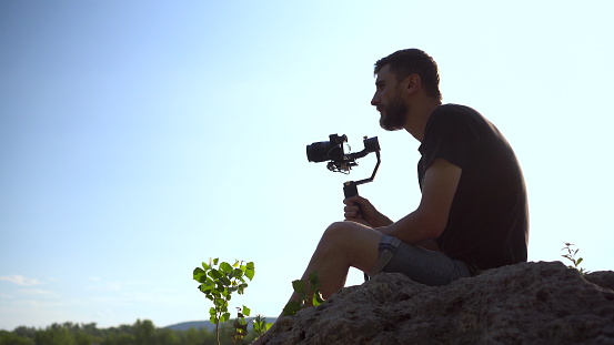 A young man sits on a stone with a camera in his hands. Man shoots at camera nature. 4k