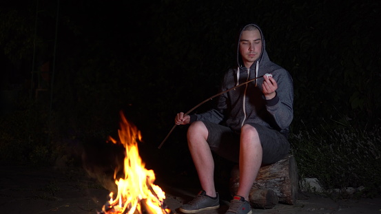 A young man in a hood fries marshmallows over a fire. Campfire at night. 4k