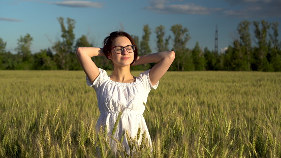 A young woman in a white dress stands in a green wheat field and touches her hair. Girl enjoys the sun in the field. 4k