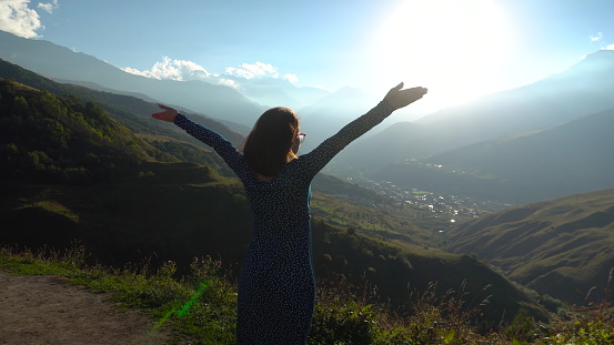 A young woman stands in a dress and raises her hands up looking at the mountains. The girl travels in the Caucasus mountains. The sun shines brightly from the side of the mountains. 4k