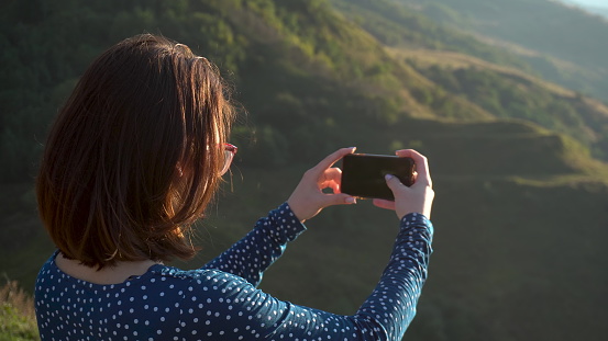 A young woman in a dress stands in the mountains and takes pictures on the phone. The girl travels in the Caucasian mountains. 4k
