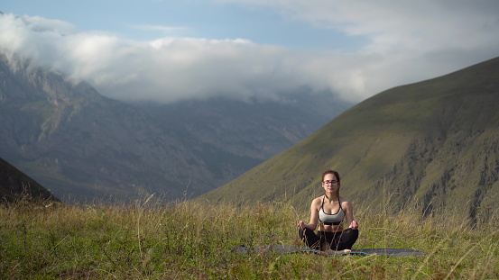 Young woman in tracksuit practices yoga performing lotus position in the mountains. 4k