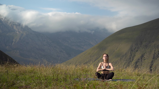 A young woman in a tracksuit practices yoga in the mountains. 4k