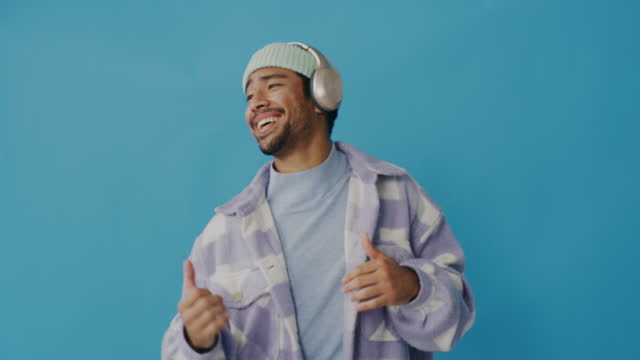 Happy asian hipster man isolated against a blue background in the studio with copyspace. Cheerful young man wearing beanie dancing to music playlist heard in bluetooth wireless headphones.