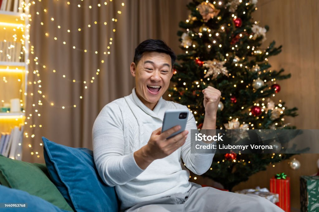 An Asian man is playing games on the phone, sitting at home near the nine-year-old Christmas tree A young Asian man is playing games on the phone, sitting at home on the couch near the nine-year-old Christmas tree. Happy with success 35-39 Years Stock Photo