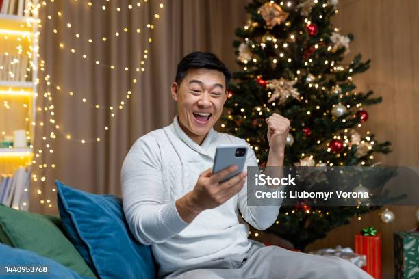 An Asian Man Is Playing Games On The Phone Sitting At Home Near The Nineyearold Christmas Tree Stock Photo - Download Image Now