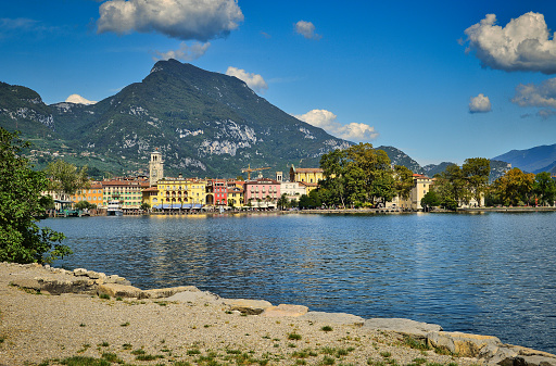 beautiful landscape of Riva del Garda, Italy- garda lake with pier, promenade, peaks, town, plants, palms, park, blue sky