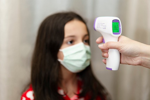 Mother taking her mask-wearing daughter's temperature with digital thermometer
