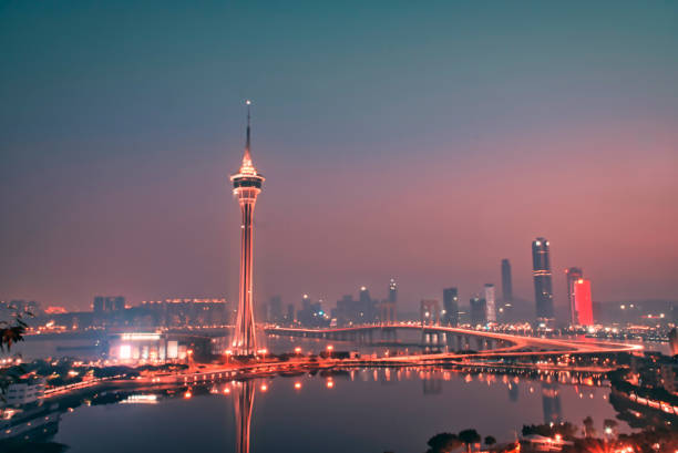 Vibrant night scene of Macau’s skyline featuring the illuminated Macau Tower, modern buildings, and a calm waterfront promenade. Colorful reflections shimmer on the water, creating a lively urban atmosphere.