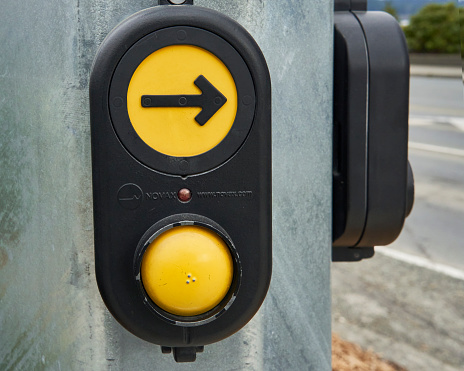 Campbell River, Canada – February 26, 2022: A closeup of the two yellow crosswalk buttons, one empty, and one with an arrow, with a black background on a pole with a glimpse of the road in front