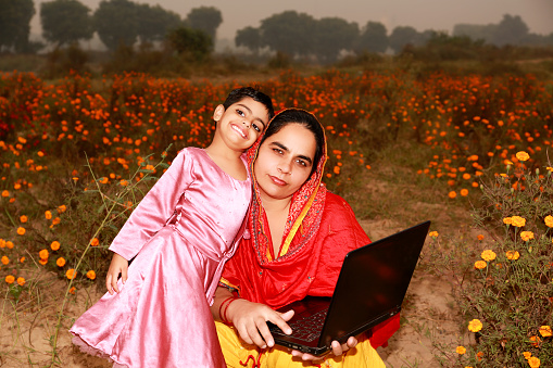 Mother and daughter of Indian ethnicity using laptop together outdoor in marigold flower field during springtime.