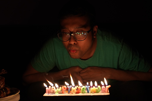 An Asian man with glasses blowing birthday cake candles