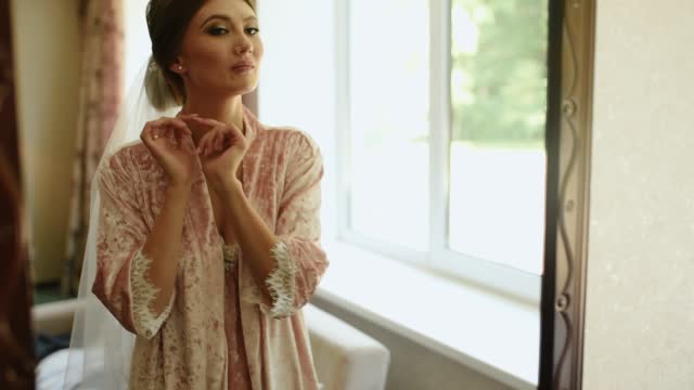 girl with veil is standing in pink nightgown and satin robe looking mirror