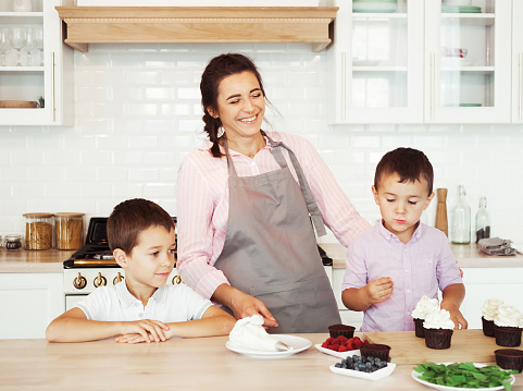 Happy family in the kitchen. Young Mom and two little boys, brothers make homemade cupcakes with berries. Family creativity, hobbies, joint activities with children.
