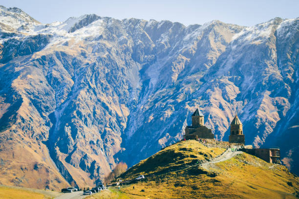 Stepantsminda, Gergeti, Georgia. Famous Gergeti Trinity Tsminda Sameba Church In Early Winter Landscape. Suv Moving Near Church In Beautiful Georgian Landscape In Early Winter