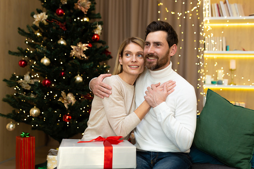 Portrait of a happy young couple, family. Young man and woman celebrating Christmas holidays at home. They sit on the sofa near the Christmas tree with gifts, hug, rejoice, look away.