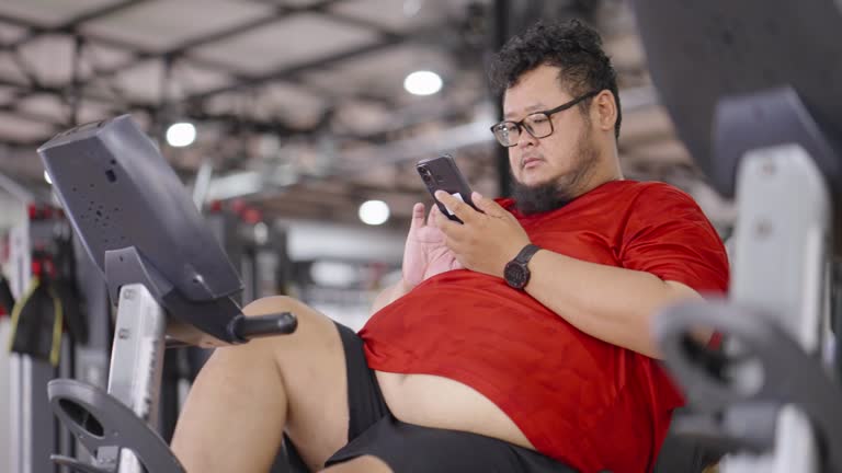 large build man using his smart phone while cycling a recumbent bike machine.