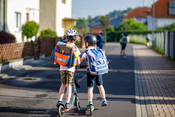 Dois meninos da escola em capacete de segurança andando com scooter na cidade com mochila em dia ensolarado. Crianças felizes em roupas coloridas andando de bicicleta a caminho da escola. Maneira segura para as crianças ao ar livre para a escola - foto de acervo