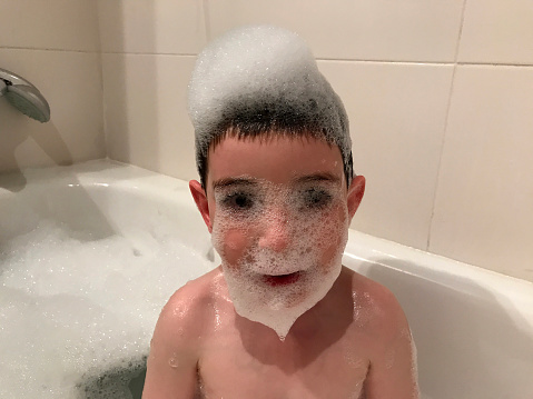 Three-year-old boy in a foam bathtub with a face full of foam. Playing and relaxing. Still looking at the camera. with foam all over his hair, face and chin. He is in a white bathtub.