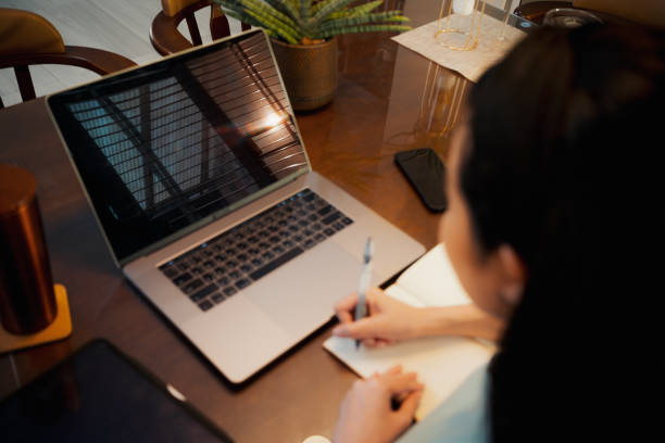 istock Over the shoulder Adult Student Woman writing on notebook paper while using laptop computer 1441014341
