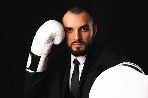 Serious young man wearing black suit and boxing gloves is ready to fight. Studio shot over black background.