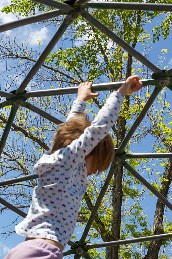A young, white, girl with brown hair hangs from bars on a metal jungle gym with a sky and tree background.  Copy space