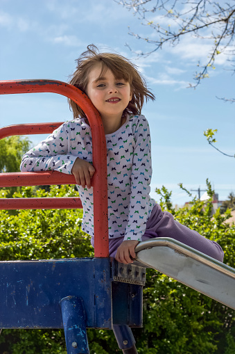 A young, white girl is seen sitting at the top of a blue and red metal slide.  She is facing the camera and appears happy.  Copy space