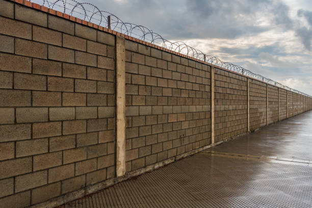 brick wall that is lost from sight along, equipped in its upper part with concertinas, wires with cutting blades as an obstacle to cross it. - razor wire imagens e fotografias de stock