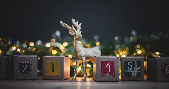 Beautiful kraft boxes advent calendar with numbers 2,3,4,5, on a wooden table with christmas deer and a burning garland on a blurred dark background, side view close-up.